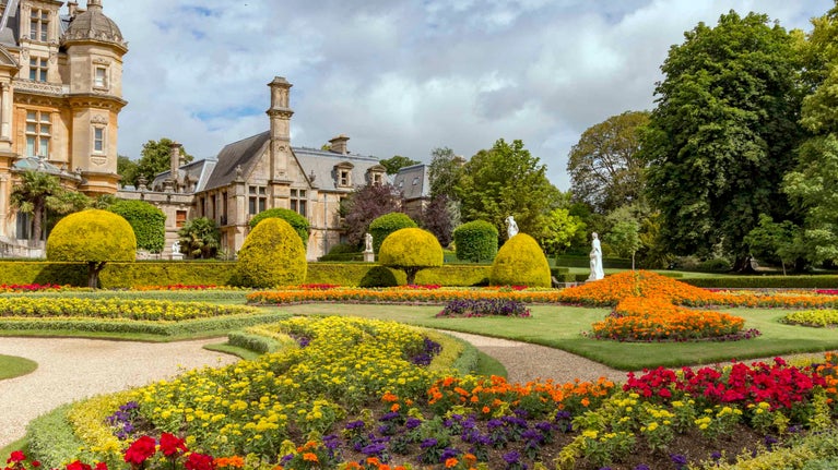 The parterre in summer at Waddesdon Manor, Buckinghamshire. Exterior view of the manor with decorative trees and flowerbeds filled with colourful flowers in the foreground.